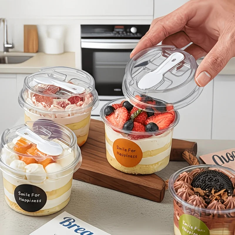 A hand holds a clear round dessert cup with a lid and spoon, filled with layered dessert, on a wooden board.