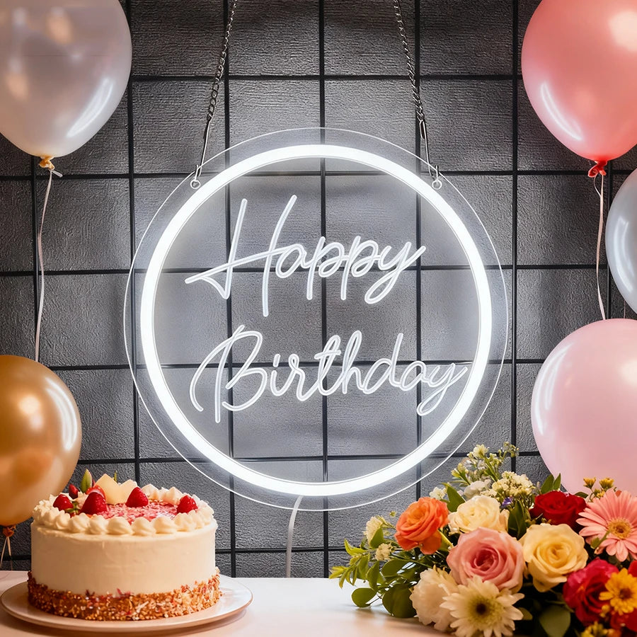 A glowing neon sign shaped like Happy Birthday is displayed on a dark wall surrounded by pink and gold balloons.