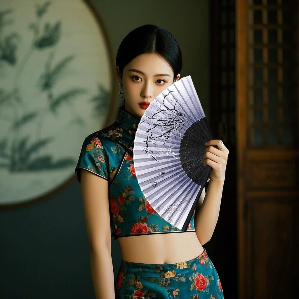 A woman wearing a teal floral top holds an open white and black bamboo folding hand fan.