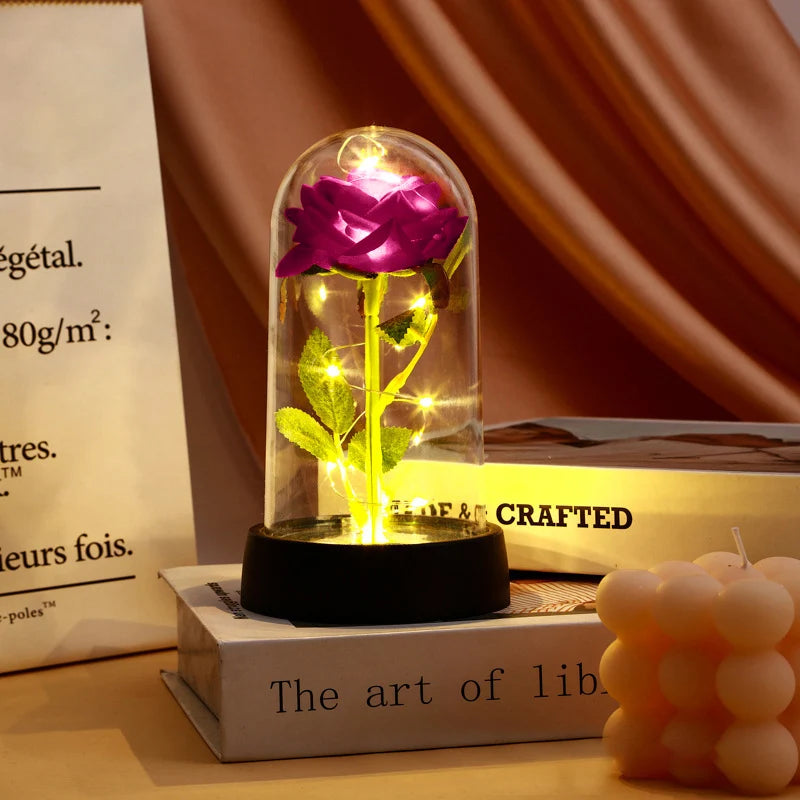 A glass dome candle featuring a pink rose and green leaves, displayed on a wooden surface.