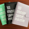 Three binders in green, black, and lilac are displayed on a wooden surface, labeled for a 100 envelope savings challenge.