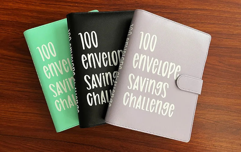 Three binders in green, black, and lilac are displayed on a wooden surface, labeled for a 100 envelope savings challenge.