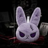 A purple fluffy bunny plush keychain with purple accents is displayed on a dark red fabric.