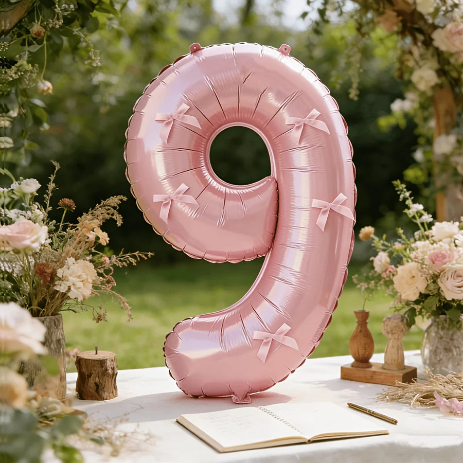 A large pink foil number balloon shaped like the number 9 with a bow, placed on a white surface surrounded by floral greenery