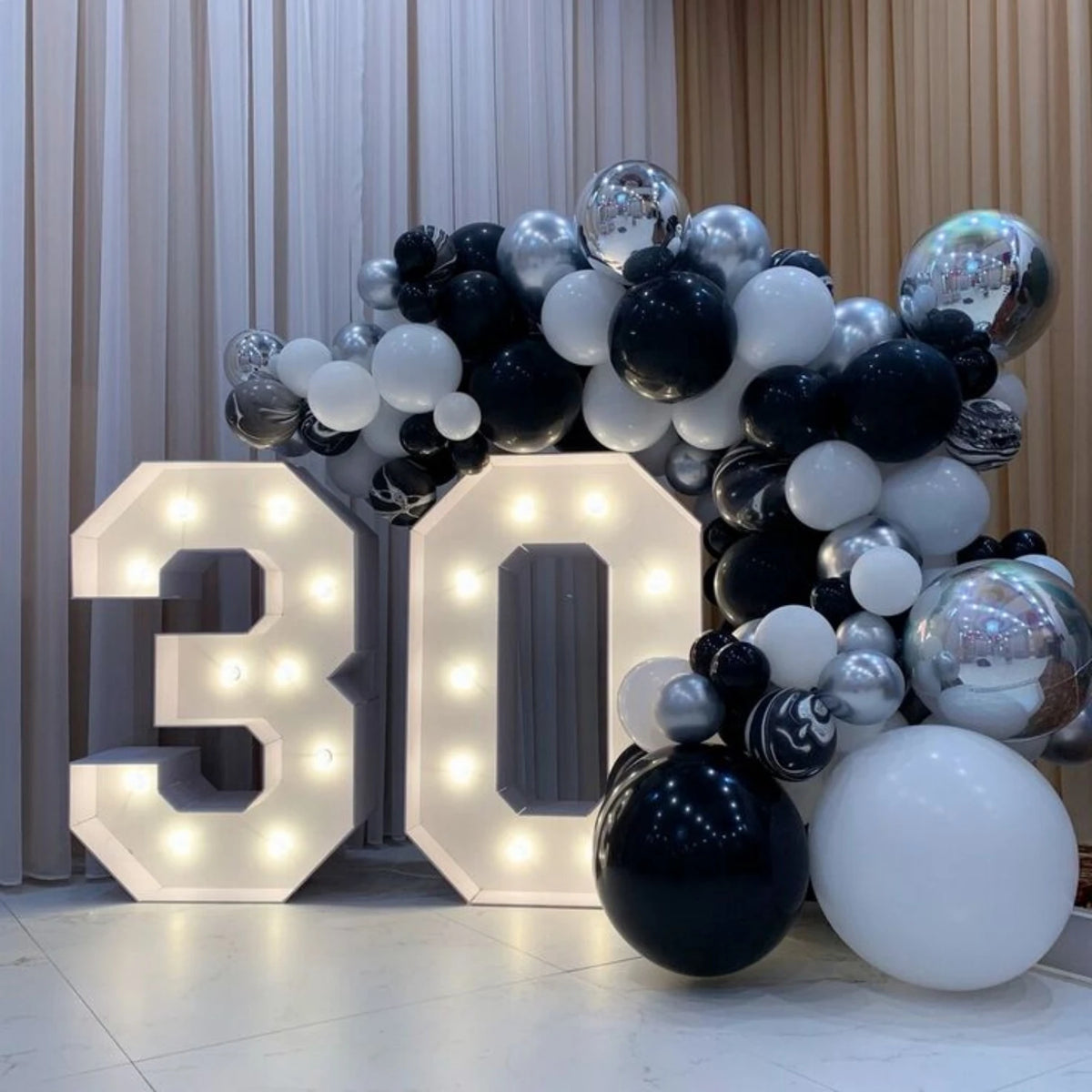 A large marquee number sign displaying 30 made of white and silver balloons, set against a backdrop of sheer curtains.