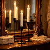 A set of tall white candles in black holders displayed on a wooden surface with a large ornate mirror.