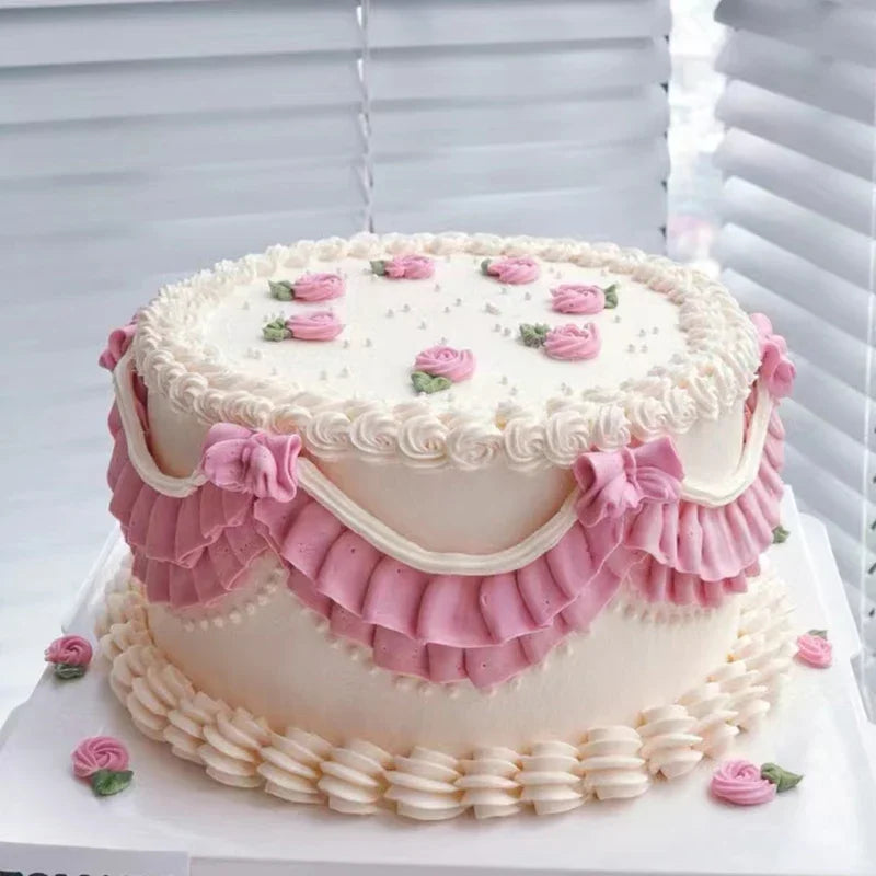 A round white cake decorated with pink floral piping and ruffled accents on a white platter.
