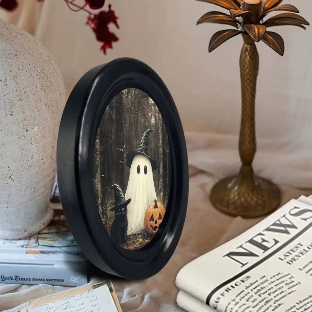 Vintage Halloween ghost and black cat wall art displayed in a black oval frame on a table.