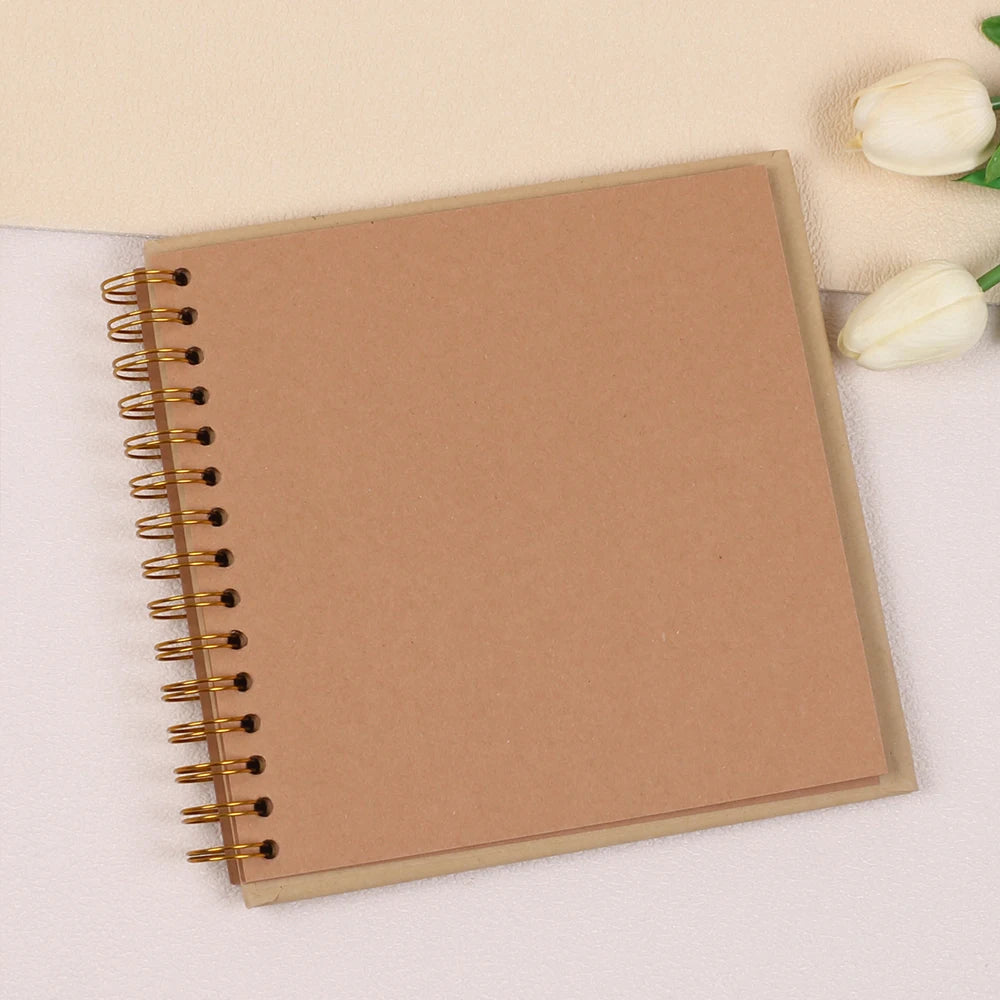 A spiral-bound kraft paper scrapbook notebook lying on a white surface with a white flower nearby.