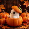 A white elf-like figure is placed inside an orange pumpkin, decorated with green leaves and set on a wooden surface surrounde