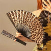 A hand holding a leopard print folding fan with a black frame against a neutral background.
