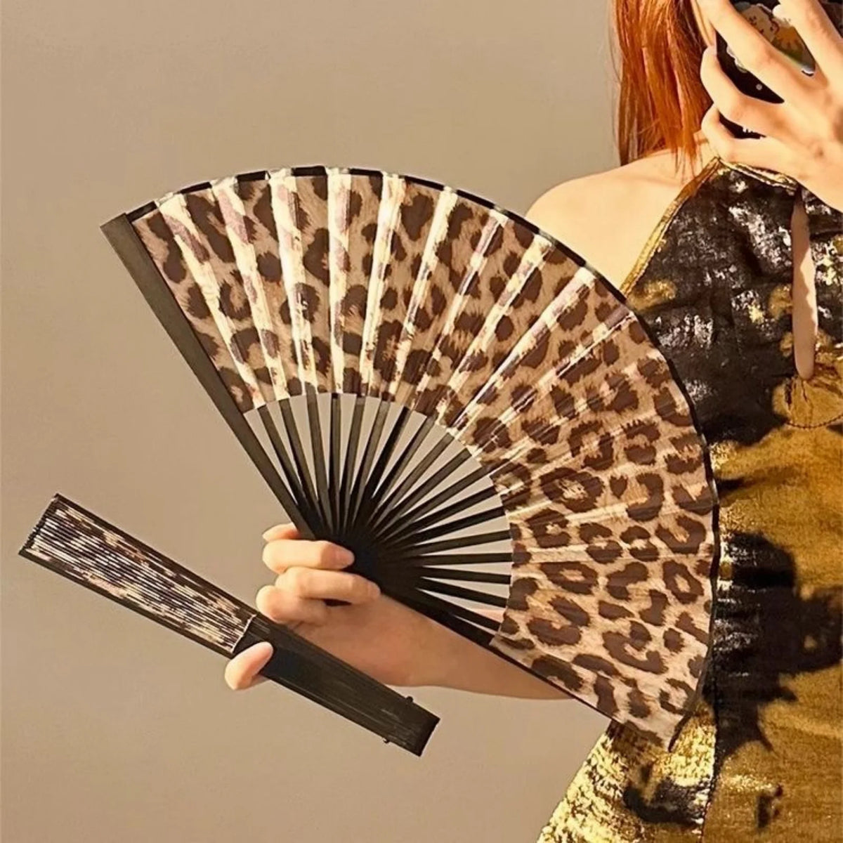 A hand holding a leopard print folding fan with a black frame against a neutral background.