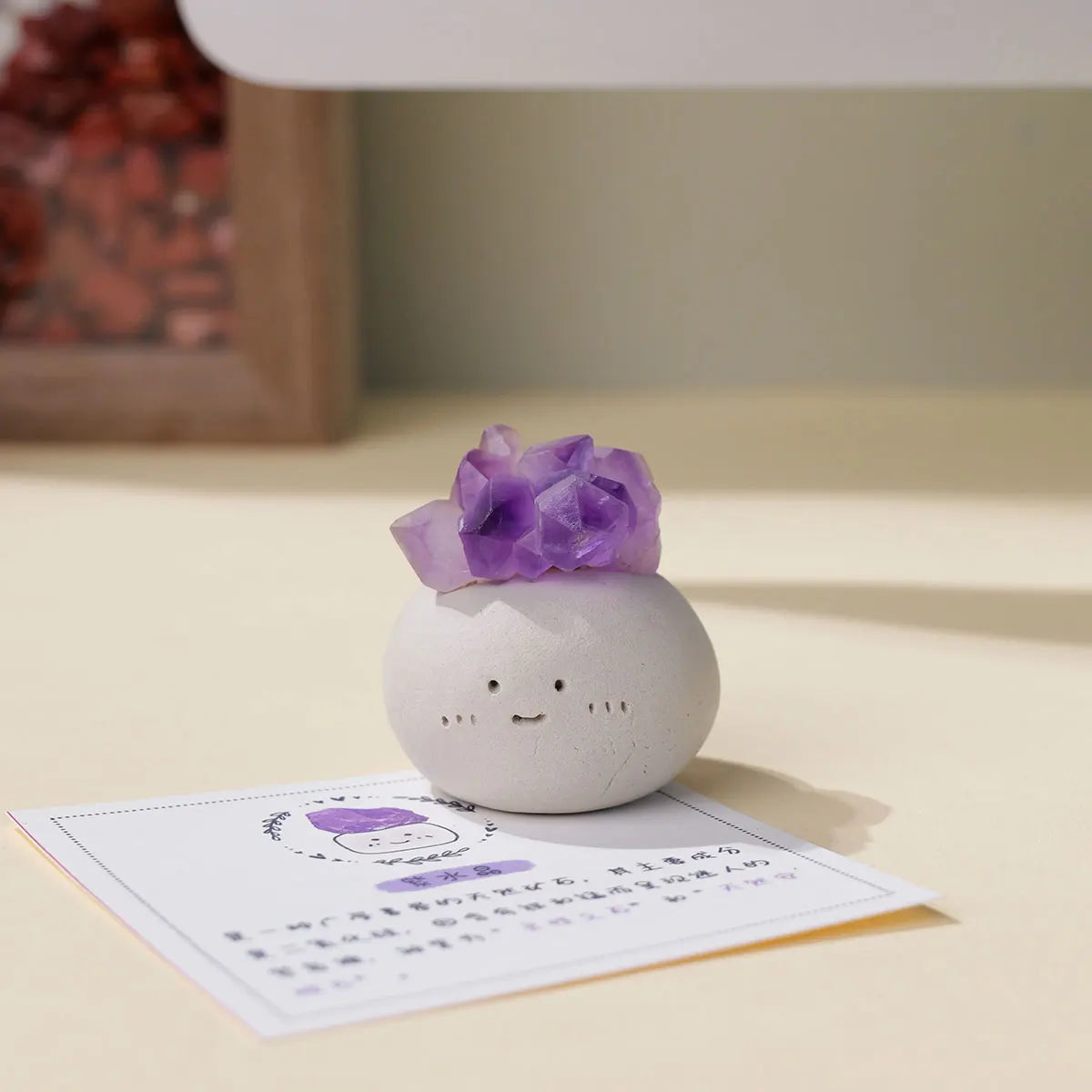 A small, smooth, white bean-shaped decorative stone with a purple crystal flower on top, resting on a card.