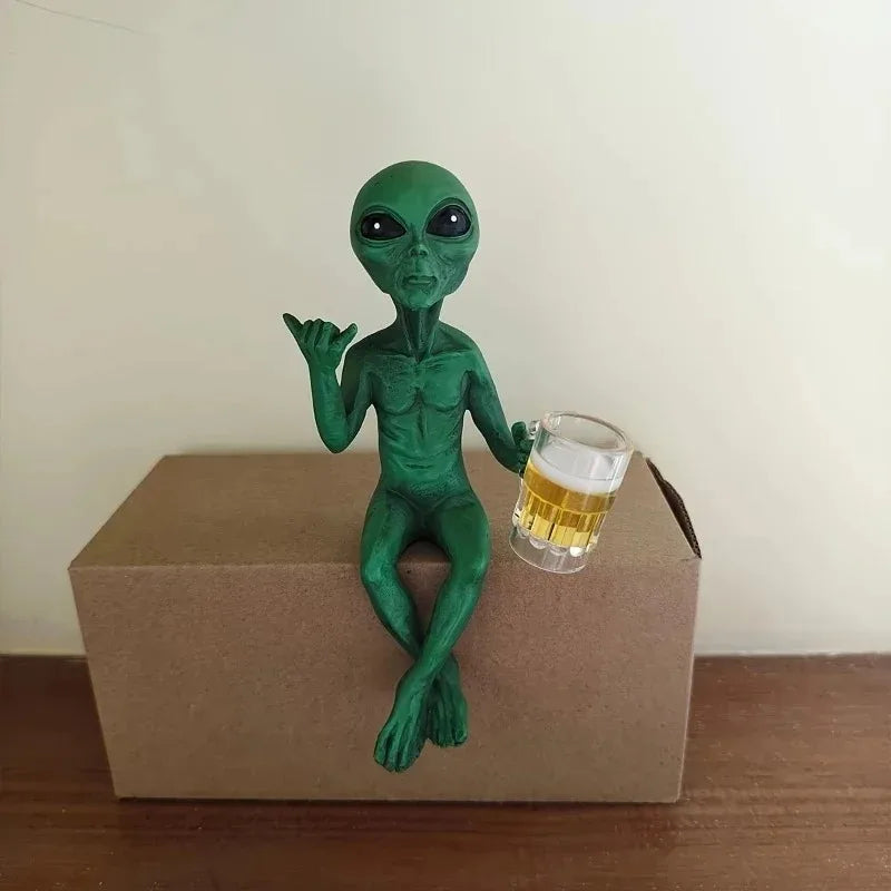Resin green alien statue holding a glass of beer on a cardboard box.