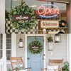 A welcome sign wreath made of artificial eucalyptus hangs on a light blue door.