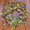 A circular wreath of artificial wildflowers featuring purple, yellow, orange, and green foliage decorated with small flowers.