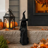 A black witch figurine with a pointed hat and cloak stands on stone steps surrounded by autumn leaves.