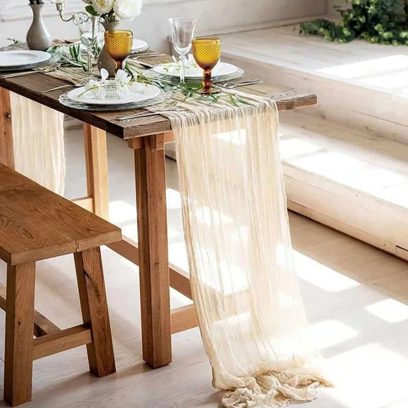 A semi-sheer beige gauze table runner is draped over a wooden table set for a meal.