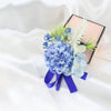 A bouquet of dusty blue silk roses and greenery tied with a royal blue ribbon, displayed next to a pink box.
