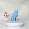 A light blue silicone mold shaped like a cat sitting on a white pedestal with pink flowers in the background.