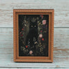 A framed painting featuring a black cat amidst dark foliage and pink flowers.