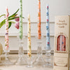 A collection of decorative printed long stick candles in pink, green, and blue are displayed on a light surface.