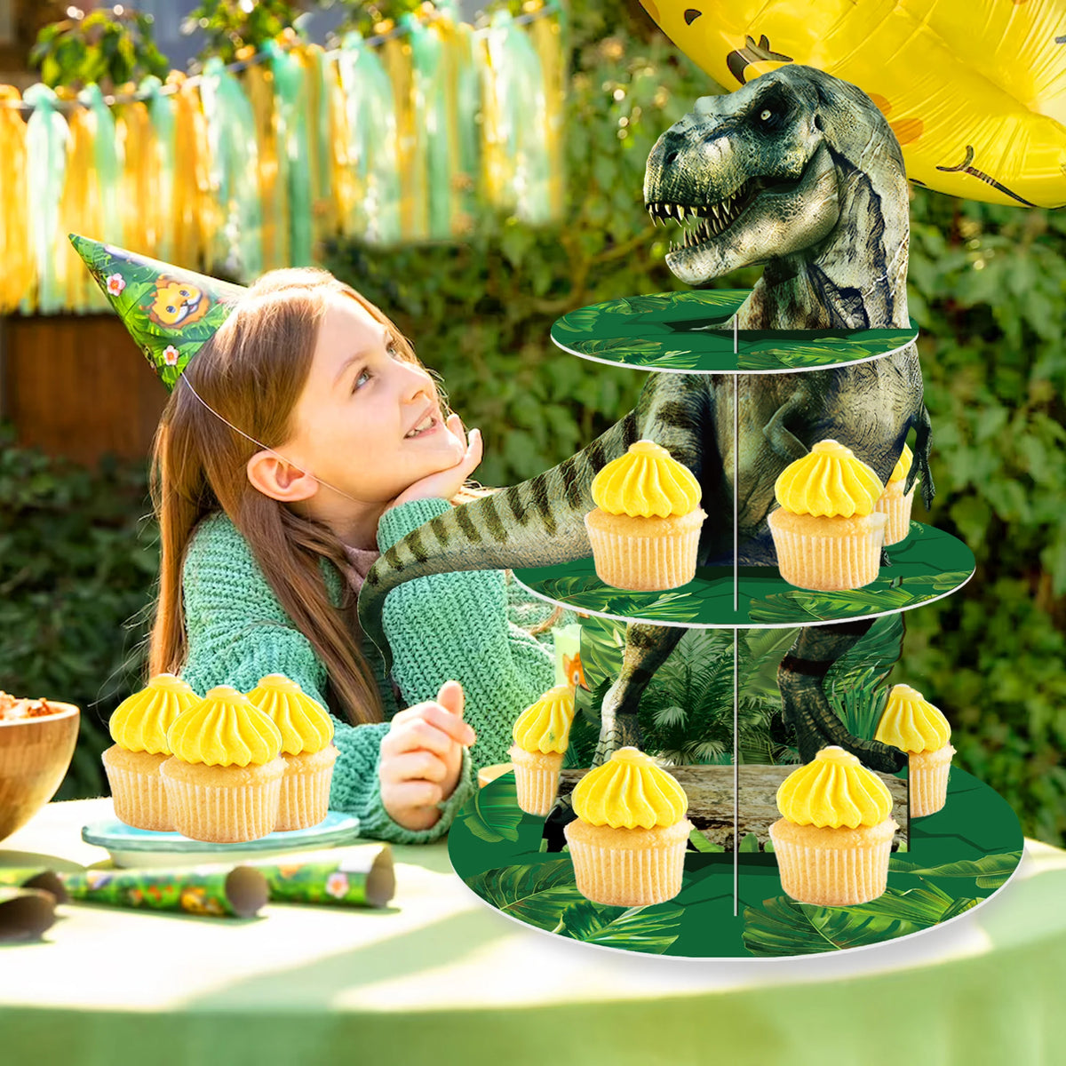A 3-tier dinosaur cupcake stand decorated with green foliage and yellow cupcakes for a party.