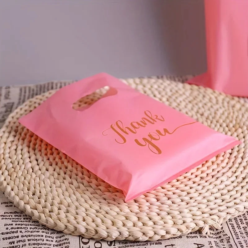A pink thank you gift bag is placed on a woven mat next to some printed paper.