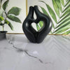 A black, sculptural heart-shaped vase sits on a white marble surface against a green leaf background.