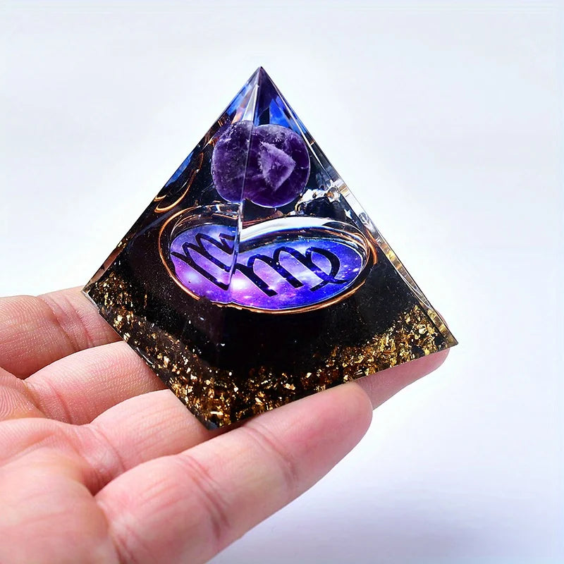 A hand holds a clear pyramid crystal with purple and gold decorative elements representing twelve zodiac signs.