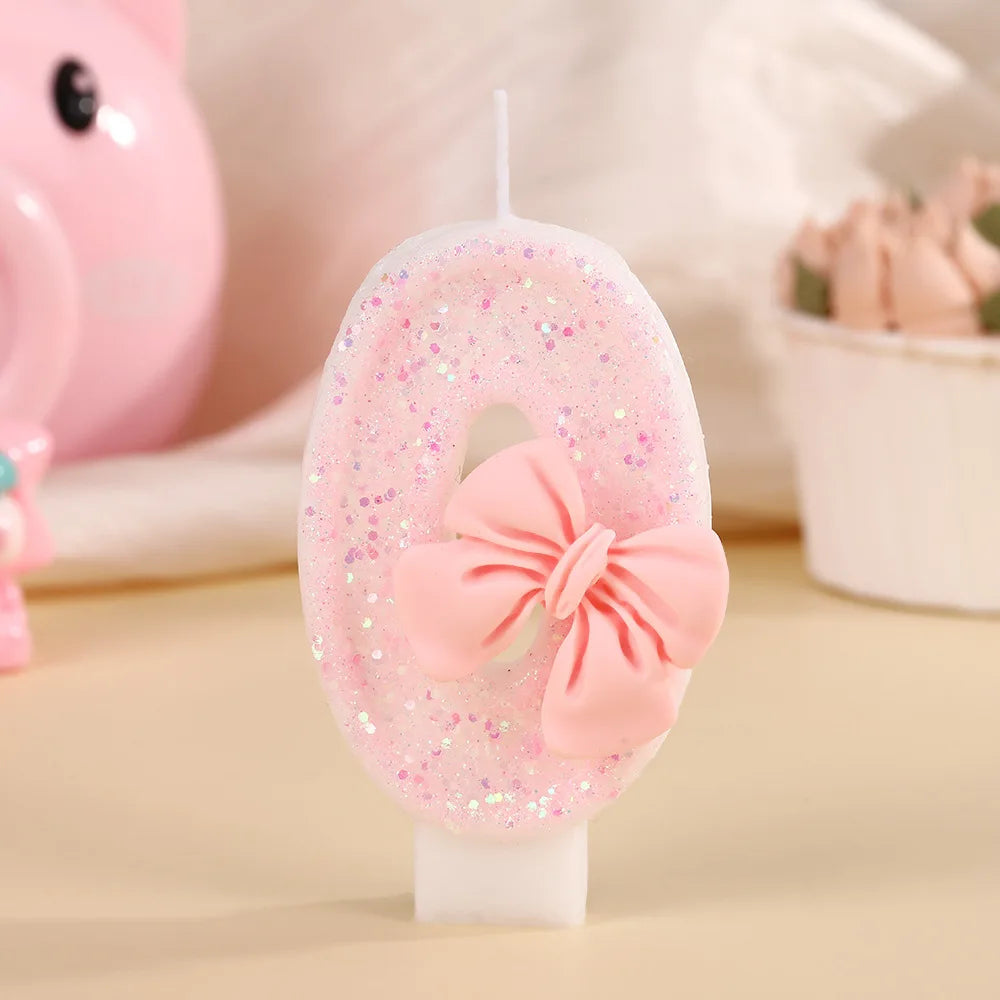 A pink bowknot birthday candle is shown on a white stand, decorated with glitter and a pink bow, next to a pink piggy toy.