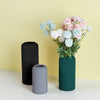 A tall dark green vase holds a bouquet of pink and blue flowers on a white surface.