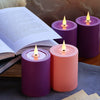 A collection of purple and coral flameless LED candles with wicks, an open book, and pine cones on a wooden surface.