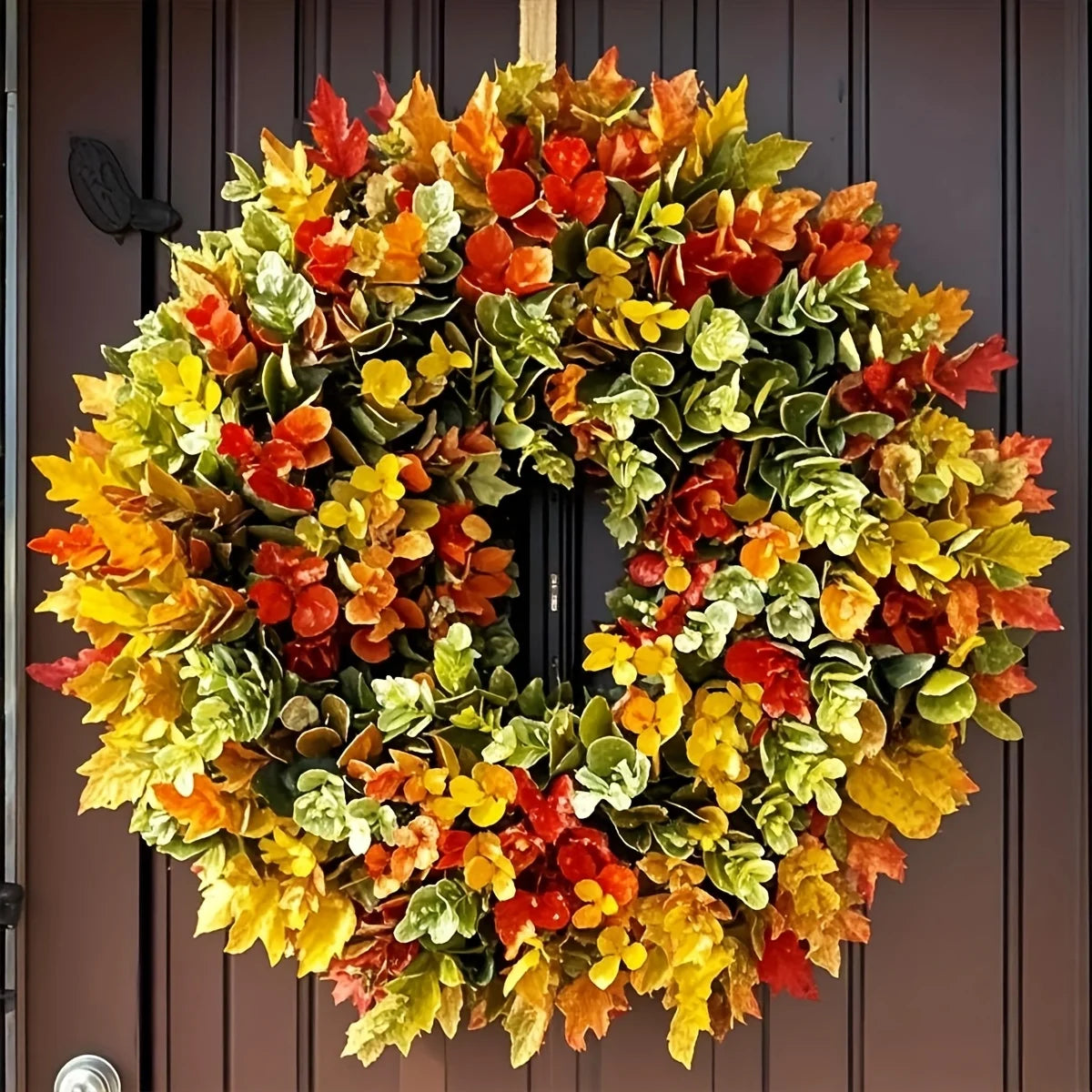 A circular wreath made of artificial autumn leaves in shades of red, orange, yellow, and green hanging on a dark wooden door.