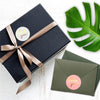 A dark navy gift box tied with a gold ribbon featuring a thank you sticker and a green monstera leaf.