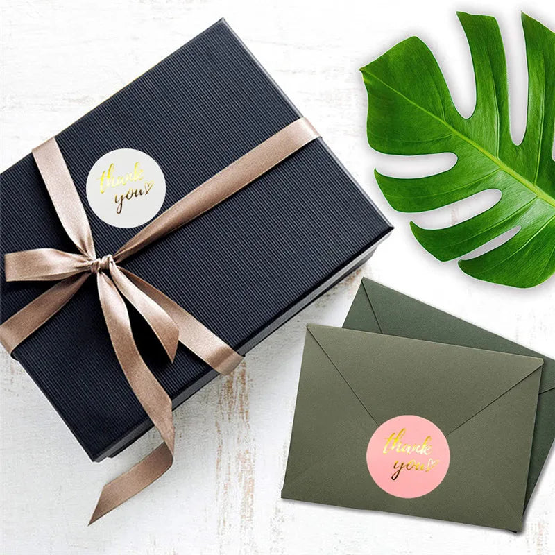 A dark navy gift box tied with a gold ribbon featuring a thank you sticker and a green monstera leaf.