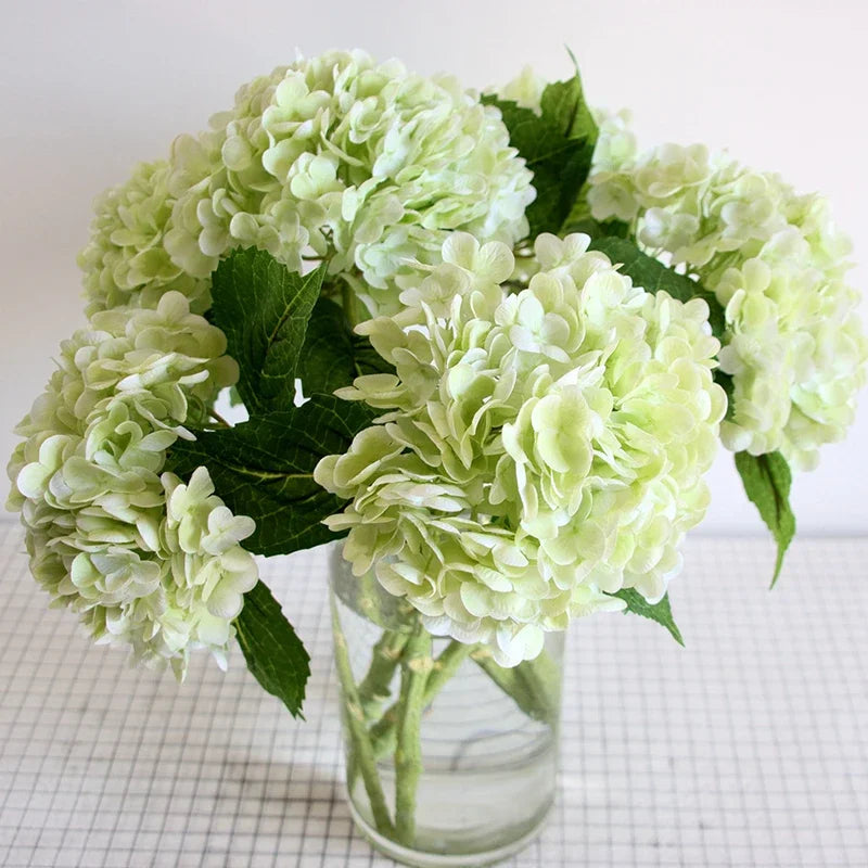 A bouquet of light green artificial hydrangea flowers in a clear glass vase.