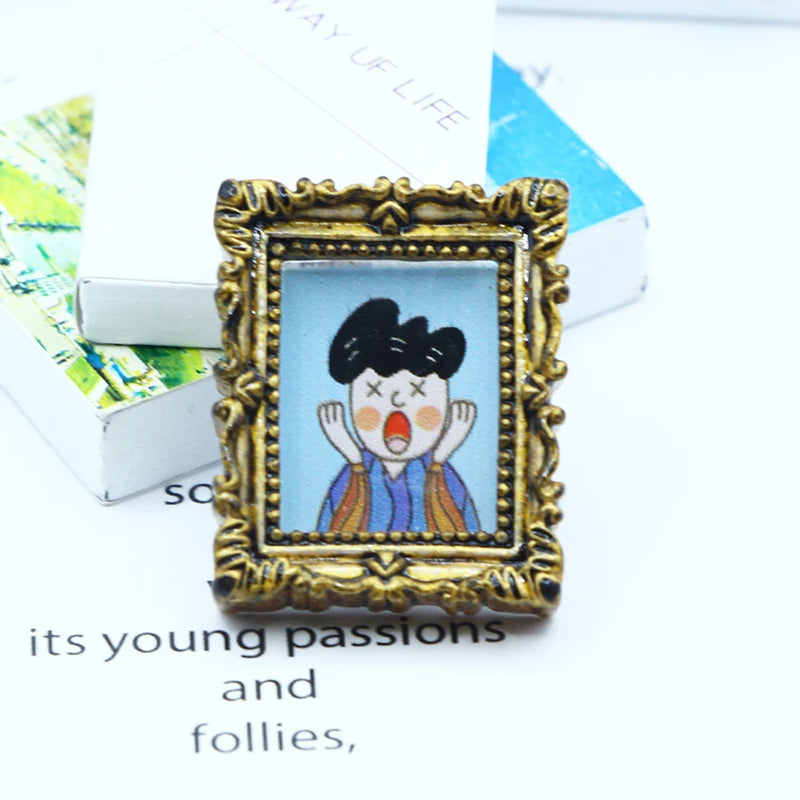 A miniature framed piece of art featuring a cartoon face with wide eyes and open mouth, set in an ornate gold frame.