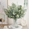 A bouquet of artificial eucalyptus leaves in various shades of green and white displayed in a glass vase.