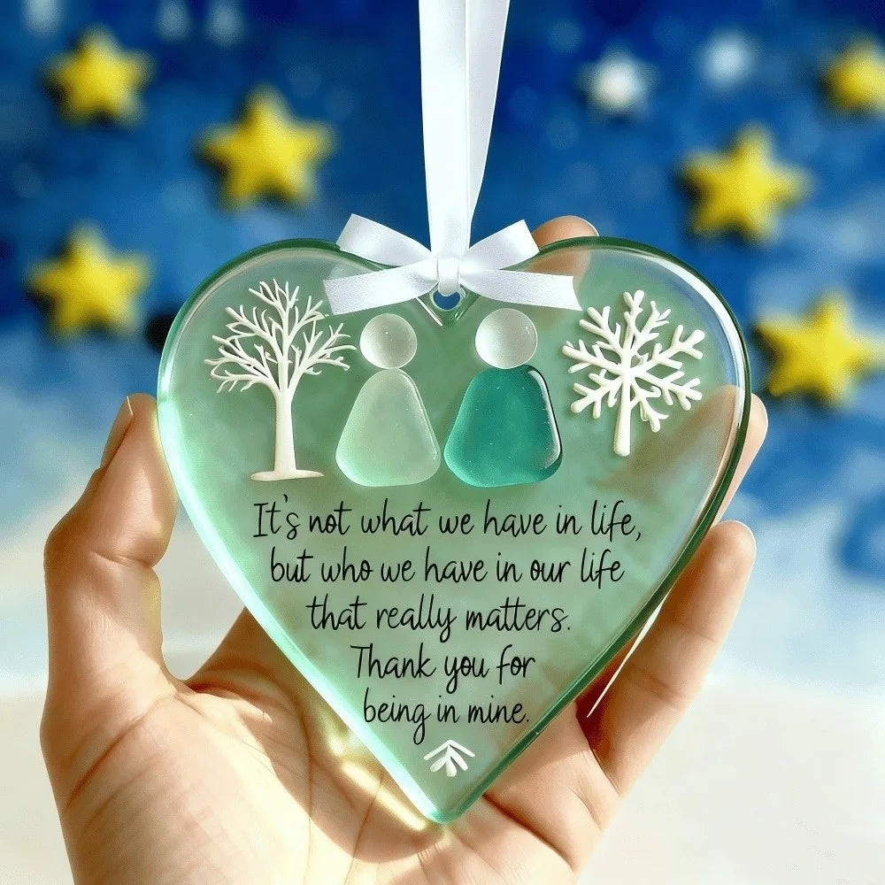 A clear green acrylic heart pendant with two silhouette figures and snowflakes is held against a blue background.