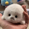 A small white seal plush toy is held in a person's hand against a blurred background.