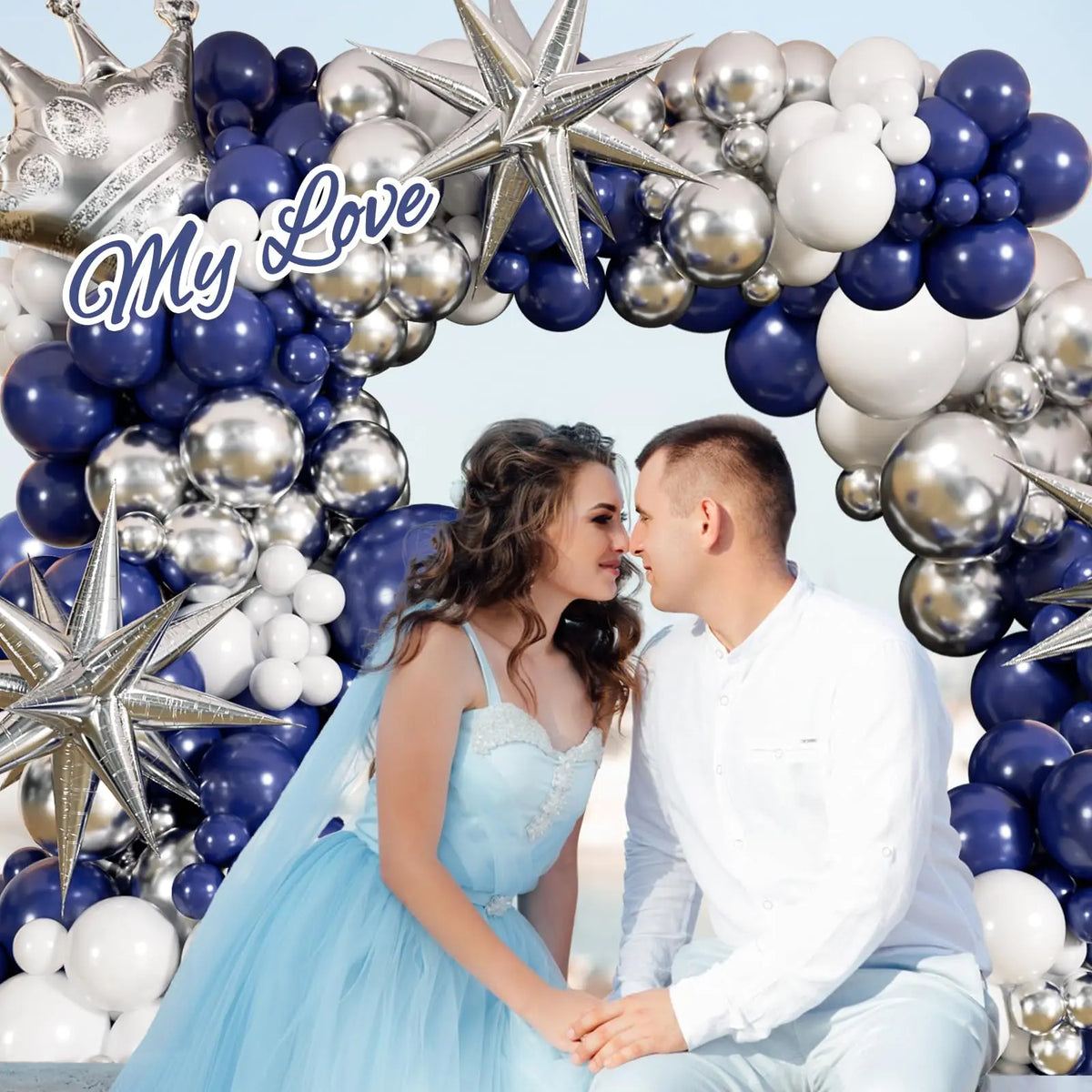 A couple is seated in front of a balloon garland arch made of navy blue and silver balloons.