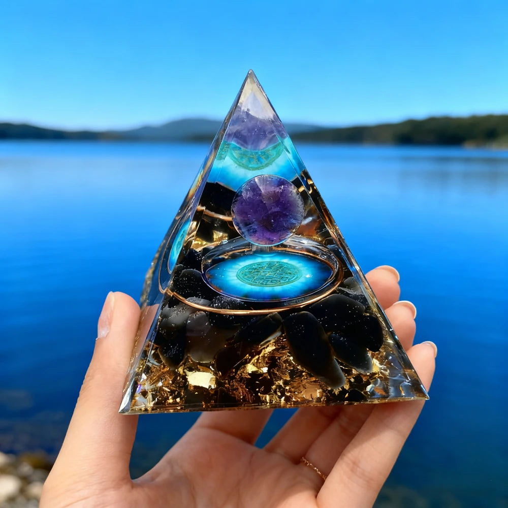 Handmade resin orgonite pyramid featuring natural obsidian and a copper coil set against a blue lake background.