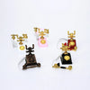 A collection of vintage rotary desk phone miniatures in gold and brass on pink, white, and black stands.