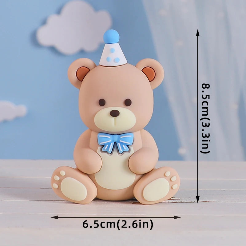 A cute, round beige bear baby block cake topper with a light blue bow and a small blue cone on top.