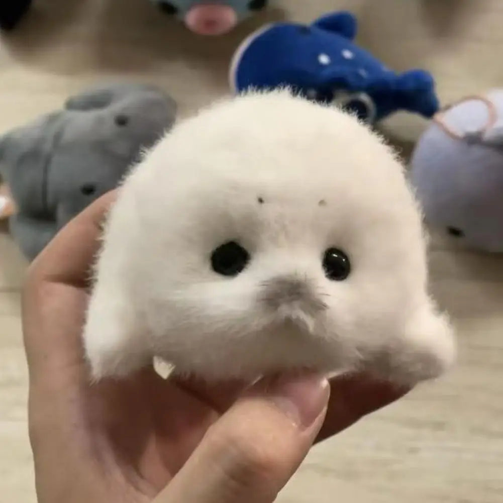 A hand is holding a small white seal plush toy against a blurred background of other blue and gray animal toys.