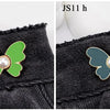 A close-up comparison of a green and a teal pearl butterfly waist tightener button on black denim jeans, labeled JS11 g and J