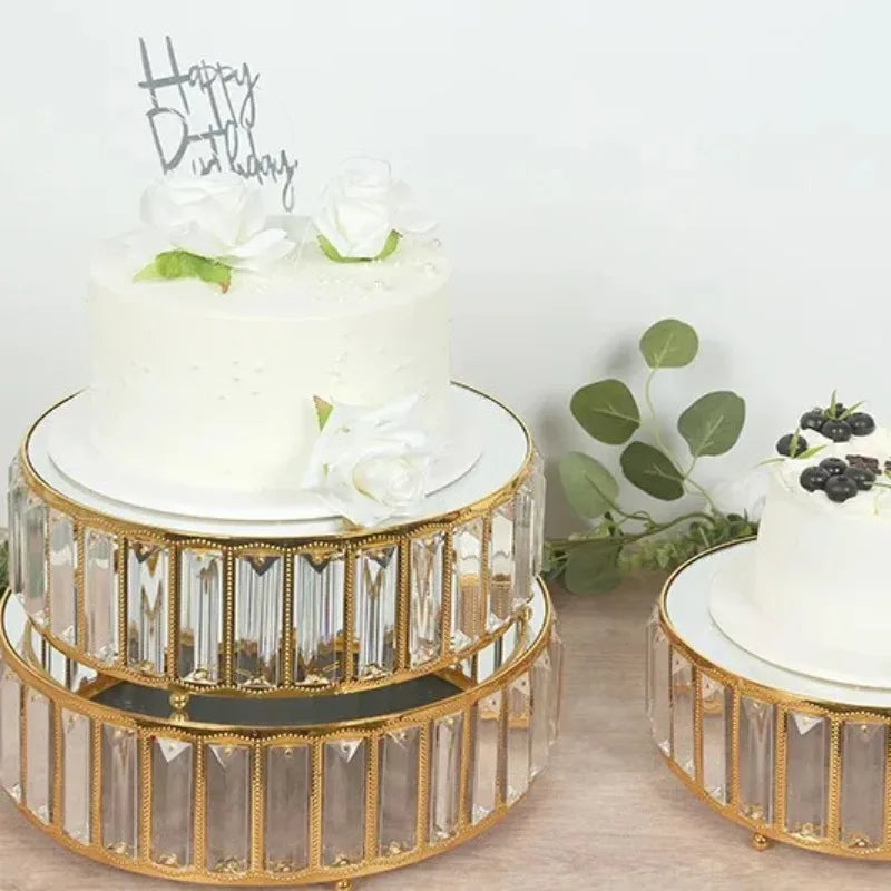 A set of three crystal and gold metal cake stands displaying a white birthday cake with flowers.