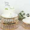 A set of three crystal and gold metal cake stands displaying a white birthday cake with flowers.
