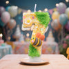 A large number seven candle decorated with green moss and gold glitter, featuring an bee design, set against a blurred birthd